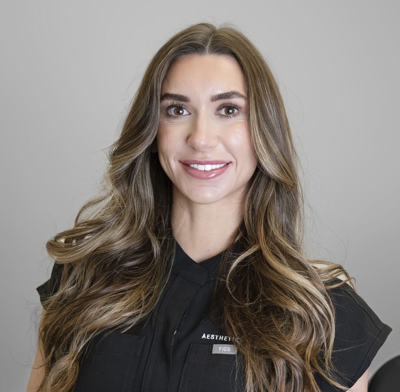 Lea Cates - SG Aesthetics & Wellness Portrait of a smiling woman with long, wavy brown hair against a gray background, wearing a black top.