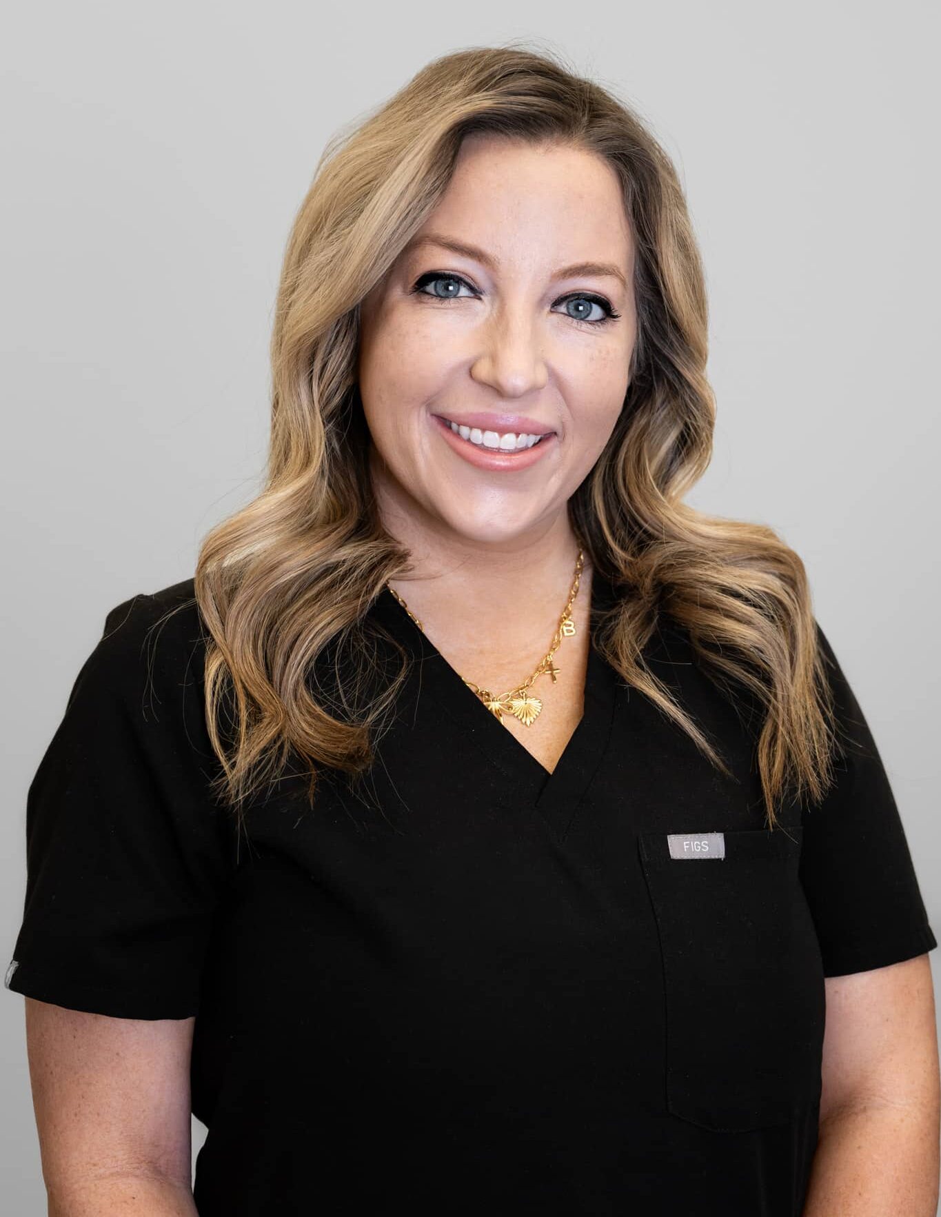 Beth Singletary - SG Aesthetics & Wellness Smiling woman with blonde, wavy hair wearing black medical scrubs and a gold necklace, head-and-shoulders portrait