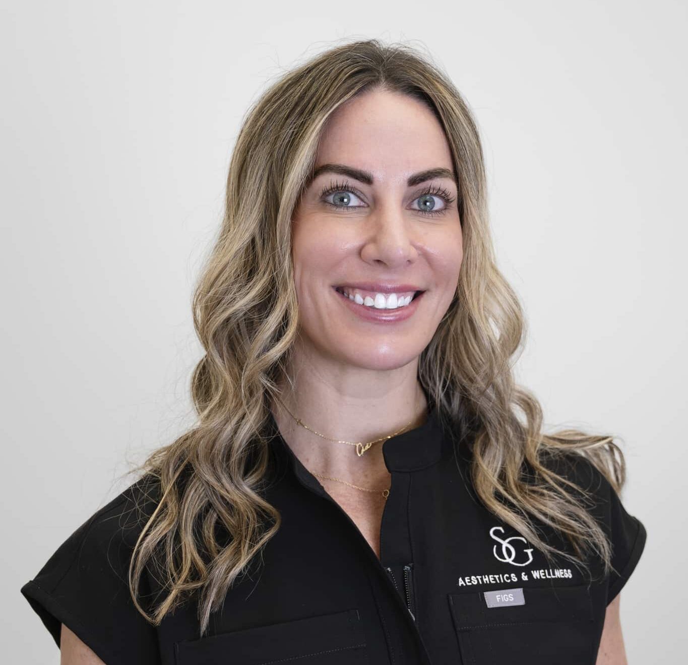 Jessica Antonelli - SG Aesthetics & Wellness Smiling woman with wavy blonde hair wearing a black uniform with an 'Aesthetics & Wellness' logo on the chest.